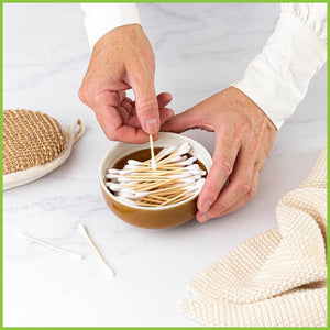 A small ceramic bowl containing bamboo cotton buds. A mans hands is holding a single cotton bud just above the others.