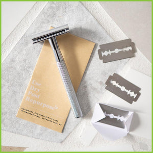 A silver safety razor lying on a 'blade return' envelope with three razor blades next to them.
