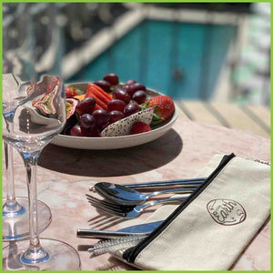 A travel cutlery set lying on a table with wine glasses and a bowl of summer fruits.
