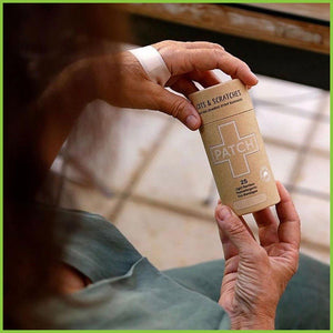 An elderly lady with a bamboo plaster on the back of her hand, holding a cardboard tube of PATCH plasters.