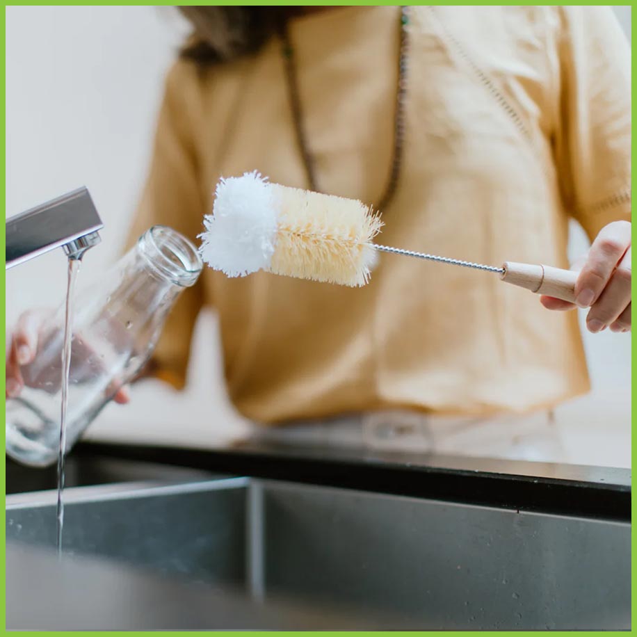 A lady stood at a kitchen sink cleaning a glass bottle. She is holding the bottle brush in one hand and the glass bottle in the other. The bottle brush is about to be put inside the glass bottle.