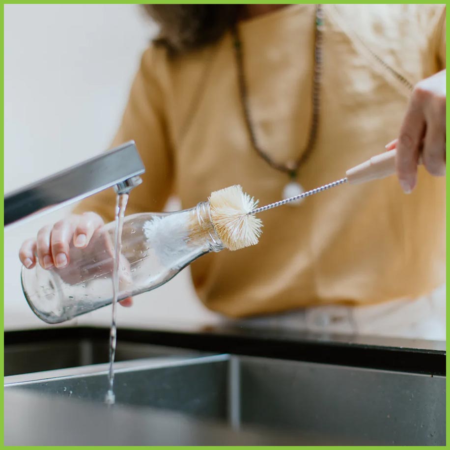 A lady stood at a kitchen sink cleaning a glass bottle. She is holding the bottle brush in one hand and the glass bottle in the other. The bottle brush is inside the glass bottle.