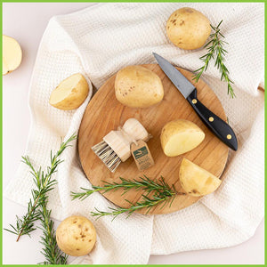 A styled photo of the veggie scrubbing brush from Go Bamboo. The veggie brush is lying on a wooden chopping board with several potatoes arranged around it. There is also a kitchen knife, sprigs of rosemary and a tea towel.