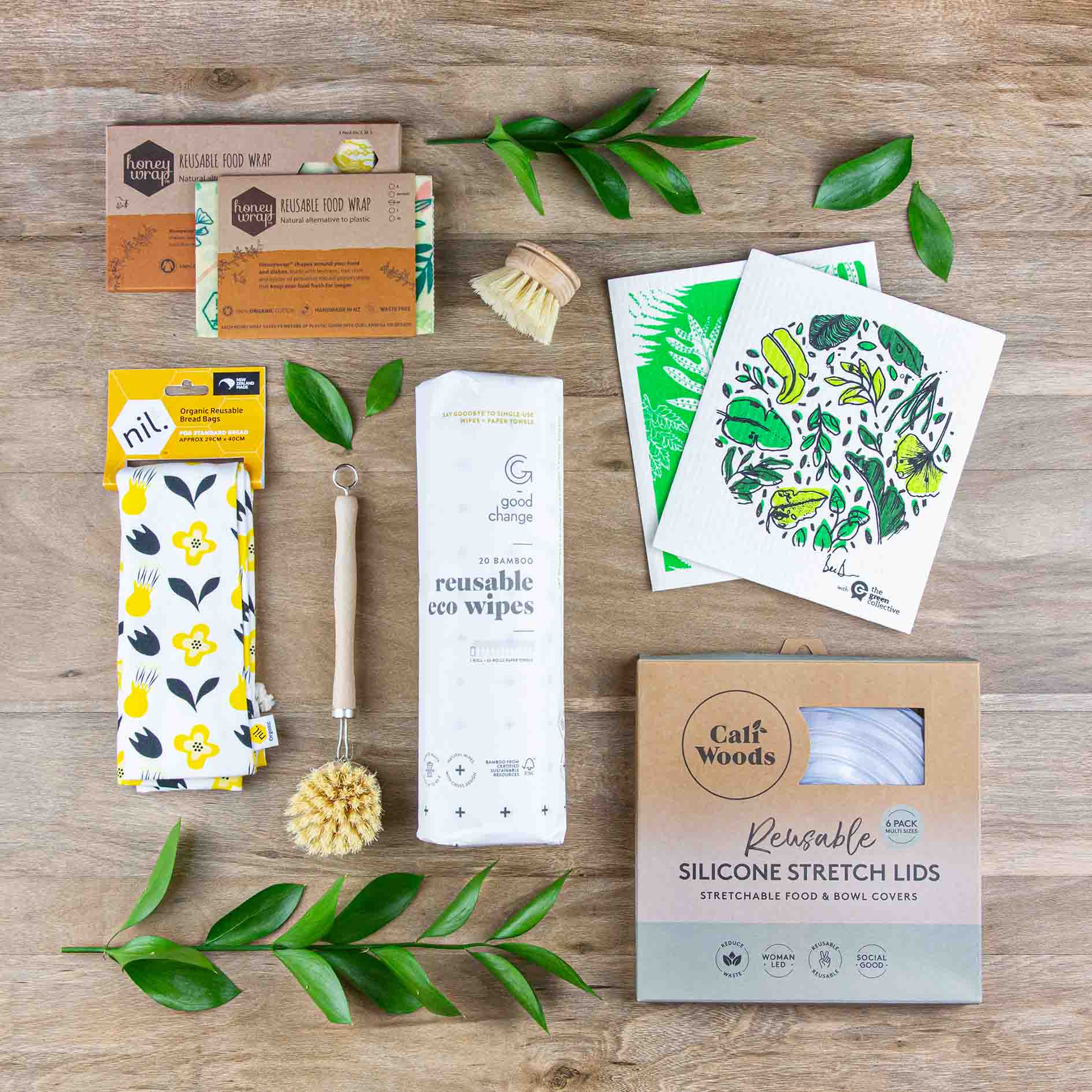 A range of zero waste cleaning and food storage products neatly laid out on a wooden floor. Products include dish cloths, beeswax wraps, eco wipes, a wooden dish brush and some silicone lids. 