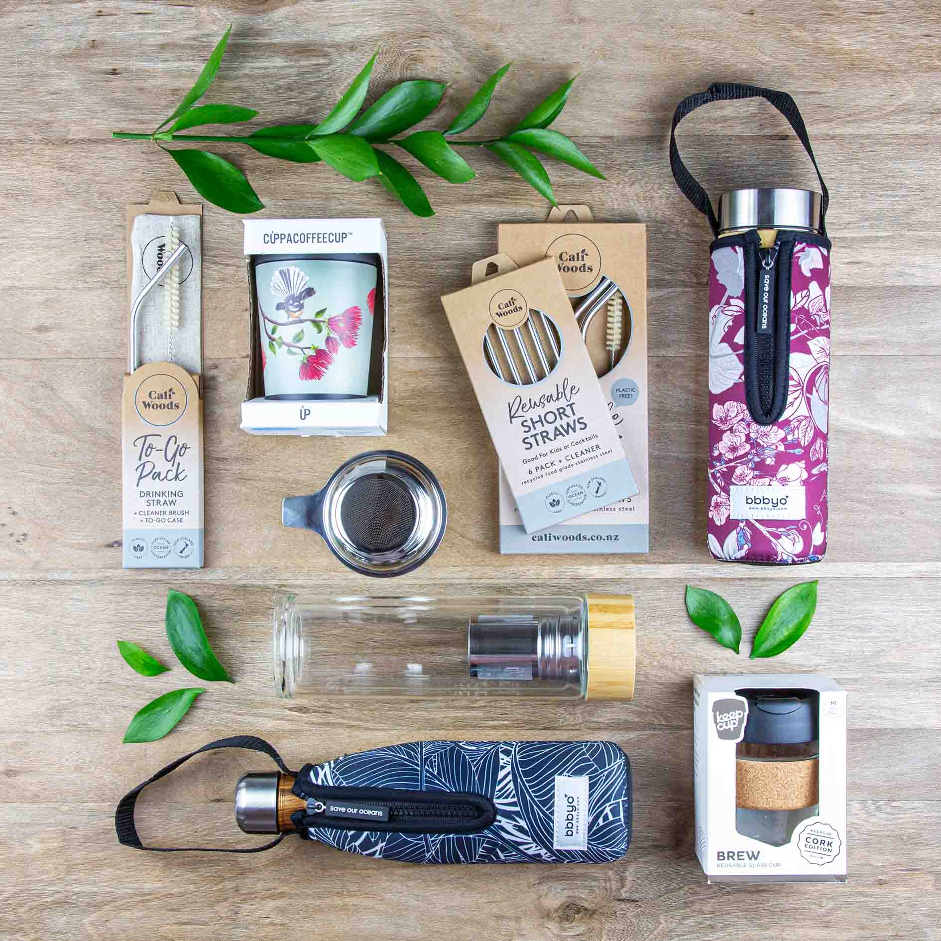 A selection of reusable drinkware such as coffee cups, water bottles, reusable straws, tea flasks and a tea infuser laid out nicely on a wooden floor.