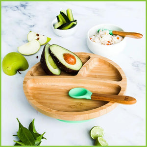 A soft silicone feeding spoon on a bamboo baby bowl. There are sliced food in and around the bowl including avocado, apple and cucumber. The baby spoon has a bamboo handle and a soft green silicone tip. 