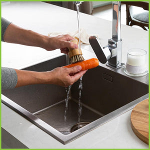 A photo of a kitchen sink with a person using a veggie brush to scrub a carrot clean under running water.