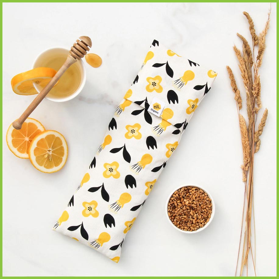 A wheat bag /heat pack, made using organic buckwheat and organic cotton. The photographed wheat bag has a white cover with bold yellow flower heads and bold black leaves on it. The wheat bag is lying flat on a marble surface with a bowl of buckwheat and a cup of honey lemon next to it.