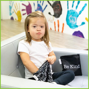 A young girl sitting on a chair eating her lunch from a 'Be Kind' Bumkins sandwich bag.