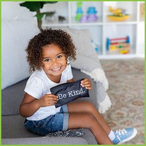 A young girl sitting on a couch smiling up at the camera. She's holding the 'Be Kind' snack bag from Bumkins.