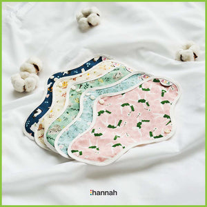 Five medium sized Hannah reusable sanitary pads lying together in a white sheet and surrounded by balls of natural cotton. Each pad has a different pattern.