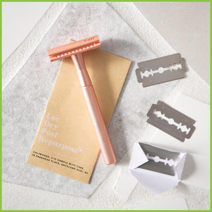 A copper safety razor lying on a blade return envelope, with three blades next to them.