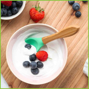 A soft silicone baby spoon in a bowl of yogurt and fruit. The feeding spoon is from the brand 'Munch' and has a bamboo handle and a soft green silicone tip.