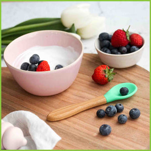 A soft silicone baby spoon laying on a table next to a bowl of yogurt and fruit. The feeding spoon is from the brand 'Munch' and has a bamboo handle and a soft silicone green tip.
