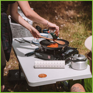 A gas stove on a camping table in the outdoors. On the table is a roll of reusable bamboo towels ready to clean up messes as needed.