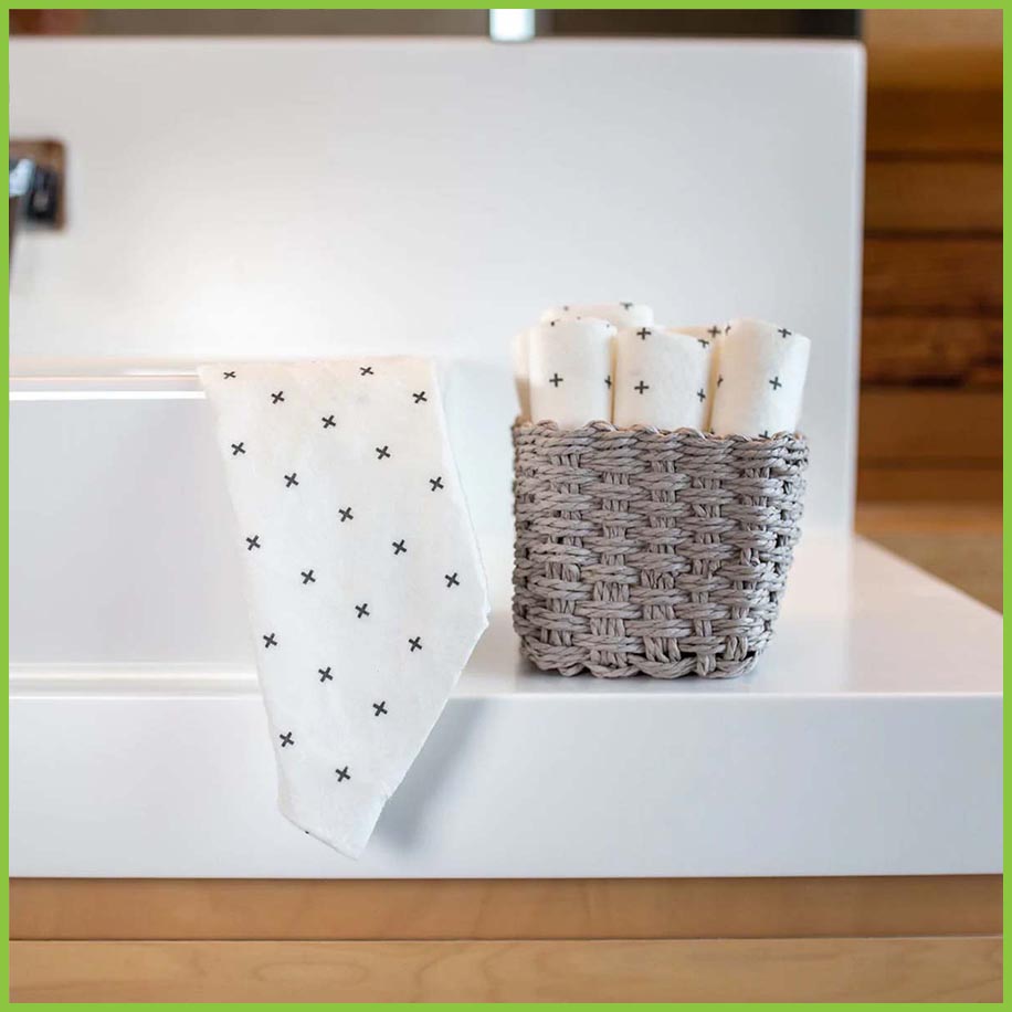 Reusable bamboo towels have been individually rolled and placed in to a small wicker basket next to a bathroom sink. One towel has been draped over the sink.