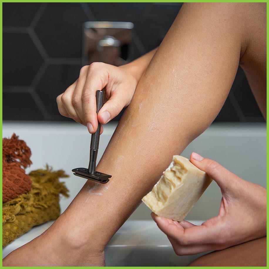A woman shaving her leg using a slate grey safety razor and a bar of shaving soap.