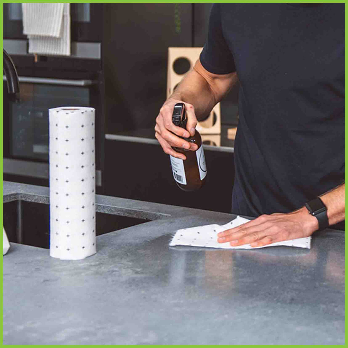 A reusable bamboo towel is being used to clean the kitchen bench. A man is in the kitchen with a cleaning spray bottle in one hand and the cleaning cloth in the other.