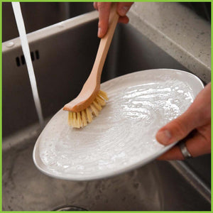 A wooden dish brush being used to scrub clean a plate in a kitchen sink.